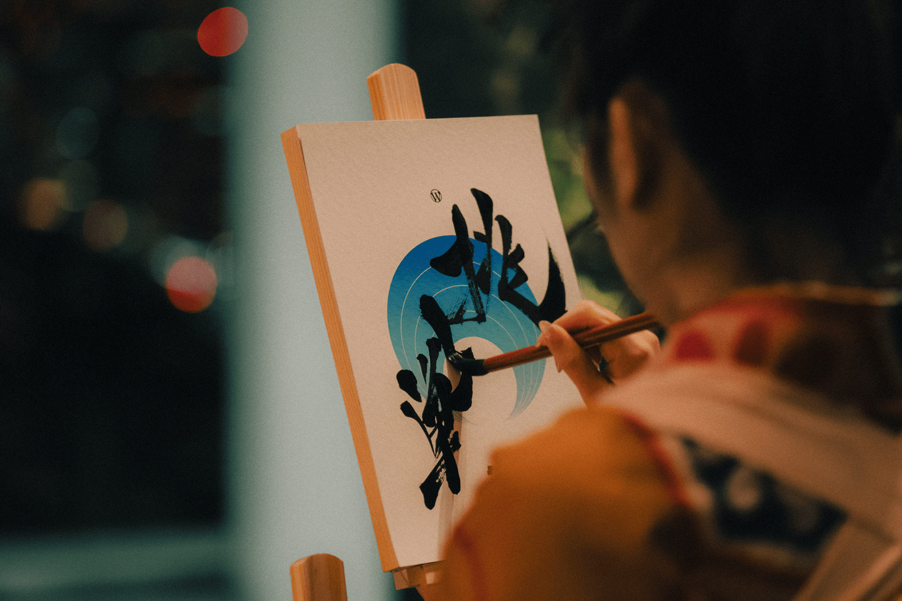 A person holding a paintbrush and writing Japanese characters on a canvas mounted on an easel.
